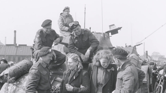 Zwart-wit-afbeelding van twee jonge blonde vrouwen die bij een tank staan, omringd door geallieerde soldaten te Groningen na de bevrijding.