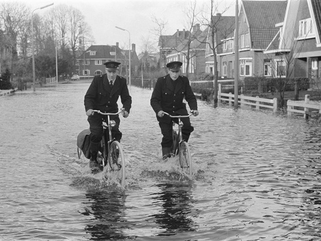 Twee postbodes van de PTT fietsen door hoogstaand water in Nijkerk in 1960